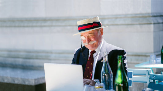 An eccentric old man in a straw boater and blazer, wearing a monocle, sitting at a laptop outdoors