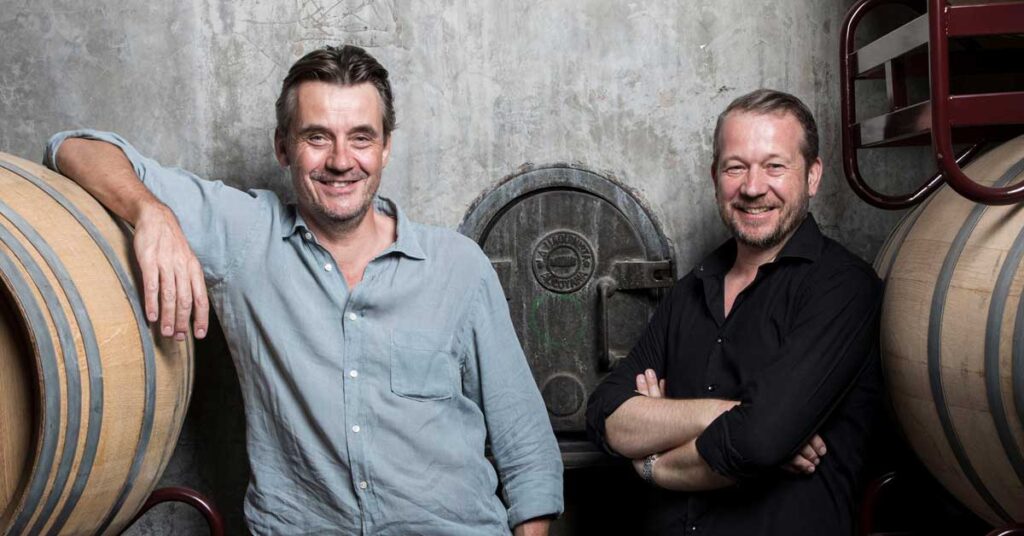 Matthew and Ben standing in a wine cellar between two large oak barrels